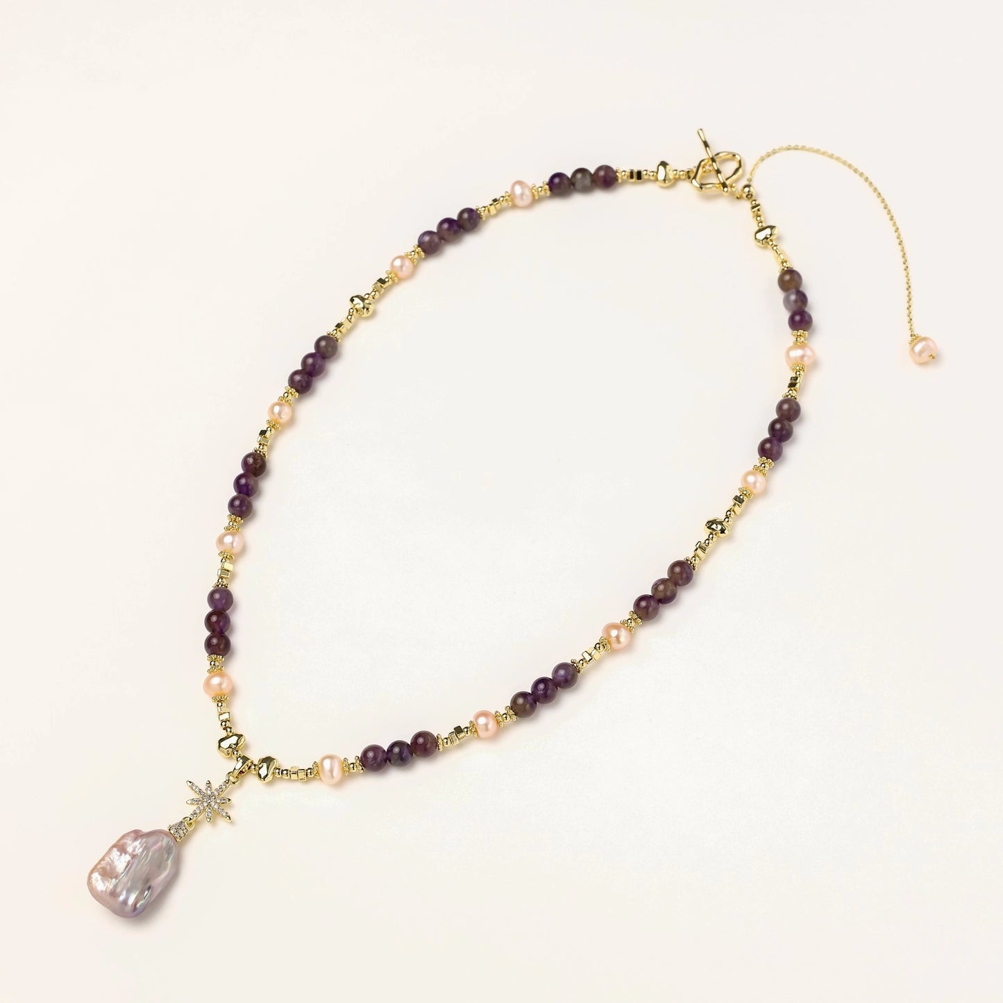 Gold necklace with iridescent baroque pearl pendant, purple amethyst beads, and crystal star charm on cream background