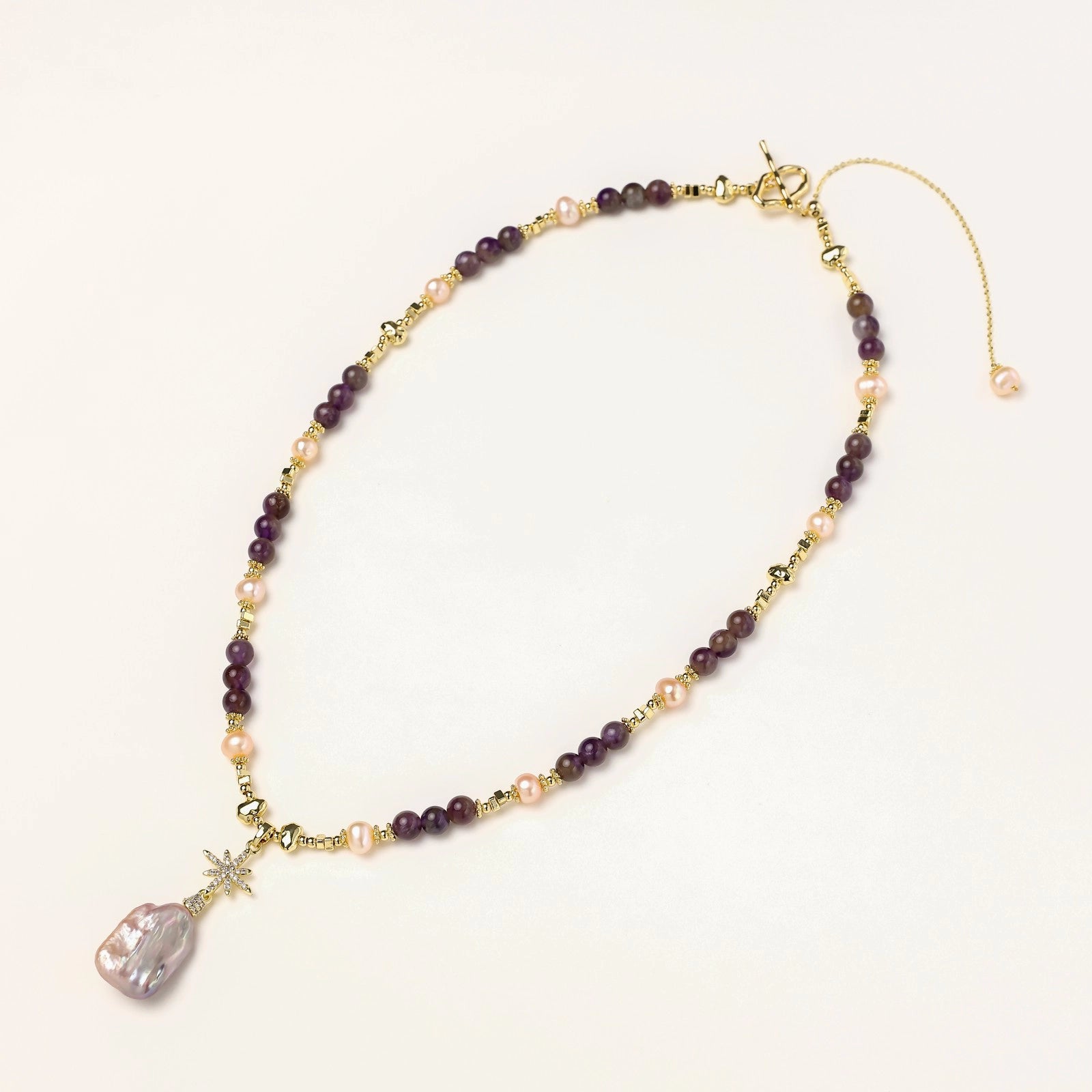 Gold necklace with iridescent baroque pearl pendant, purple amethyst beads, and crystal star charm on cream background