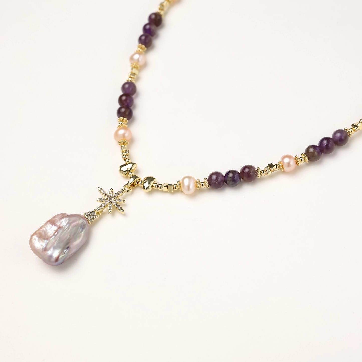 Gold necklace with iridescent baroque pearl pendant, purple amethyst beads, and crystal star charm on cream background