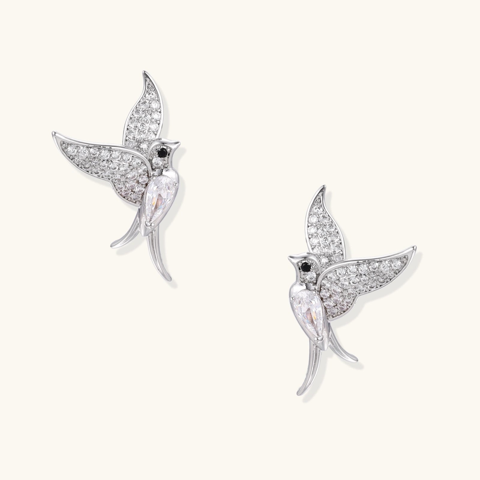 Pair of white gold-plated hummingbird stud earrings with pavé zirconia stones and teardrop crystal bodies on cream background
