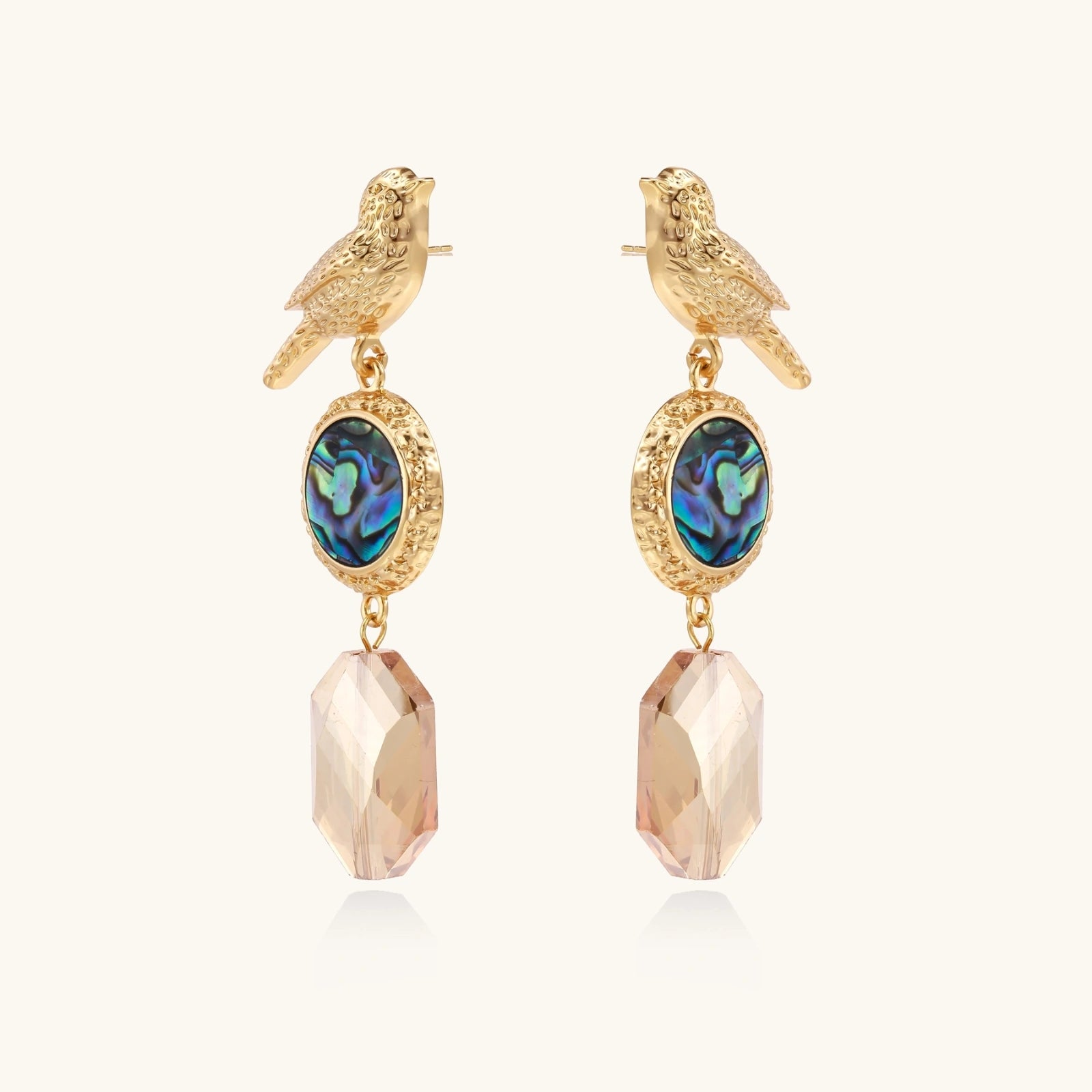 Gold-plated bird stud earrings with iridescent abalone shell medallions and champagne crystal drops on cream background