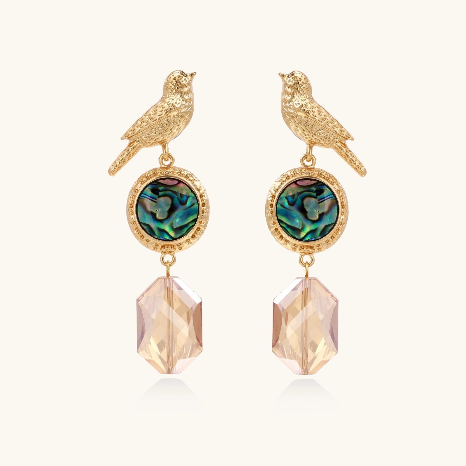 Gold-plated bird stud earrings with iridescent abalone shell medallions and champagne crystal drops on cream background