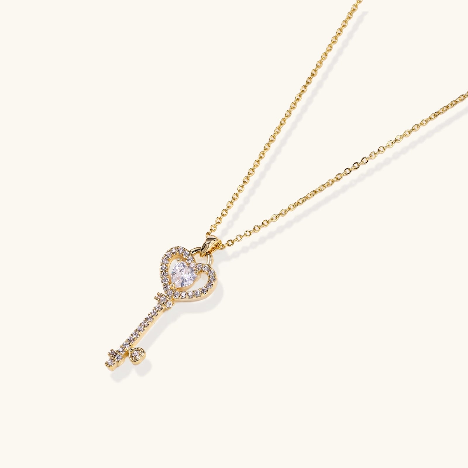 Delicate gold key pendant with heart-shaped bow and crystal pavé detailing on fine chain against cream background.